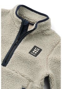 Fleece bunda v svetlej béžovej farbe s námorníckym rámom, s vysokým golierom, zipsovým zapínaním a textúrovanou látkou. Obsahuje čierny logotyp.