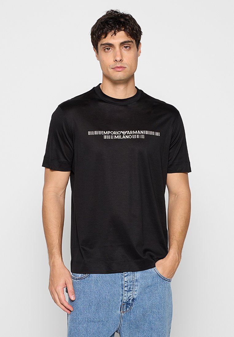 Hombre con camiseta negra Emporio Armani Milano y vaqueros azules, de pie con una mano en el bolsillo frente a un fondo liso.