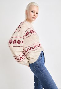 Κρέμα πουλόβερ με κόκκινους χιονανθρώπους και γεωμετρικά μοτίβα, υπερμεγέθης εφαρμογή, ριγωτοί μανσέτες, φορεμένο με μπλε τζιν ψηλής μέσης.