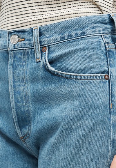 Κοντινό πλάνο ανοιχτό μπλε τζιν με μεταλλικό κουμπί και νίκελ, φορεμένο με μπεζ και μαύρη ριγέ μπλούζα.