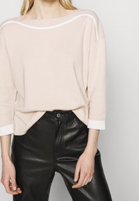 Femme portant un haut beige à manches longues avec des bordures blanches et un pantalon en cuir noir taille haute, se tenant devant un fond uni.