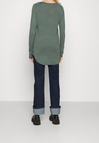 Grünes Langarmshirt mit abgerundetem Saum und enganliegenden Ärmeln, kombiniert mit dunkelblauen Jeans mit aufgekrempelten Bündchen und klobigen schwarzen Schuhen.