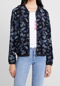 Svart bombarjacka med ett paisleymönster i blå och gröna nyanser. Ribbat manschetter och nederkant, dragkedjestängning, bärs över en vit T-shirt.