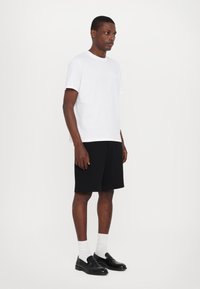 Tricou alb din bumbac cu margini roz deschis, asociat cu pantaloni scurți negri. Modelul poartă mocasini negri și șosete albe până la glezne, stând în fața unui fundal neutru.