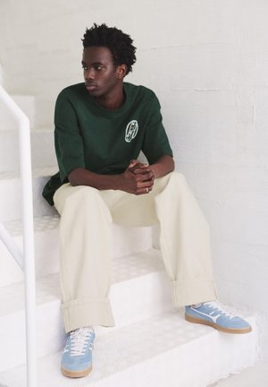 Mladi moški v zeleni srajci in bež hlačah sedi na belih stopnicah, gleda v stran s sklenjenimi rokami, nosi modre superge.