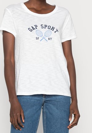 Osoba ubrana w białą koszulkę z krótkim rękawem z napisem "GAP SPORT" i grafiką skrzyżowanych rakiet tenisowych, w połączeniu z niebieskimi dżinsami.