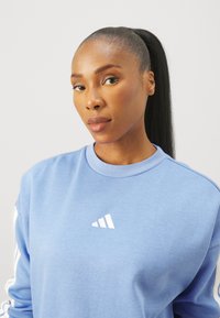 Ljusblå sweatshirt med rund halsringning och vitt logotyp på bröstet; har vita ränder längs ärmarna. Slät tygkvalitet.