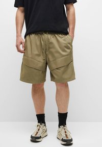 Khaki korte broek met een elastische tailleband, twee grote voorzakken en een gladde textuur. Draagt met een zwart T-shirt en sportieve schoenen.