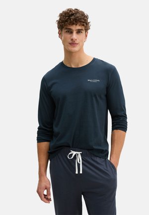 Mann mit lockigem Haar, der ein marineblaues Langarmshirt mit dem "Marc O'Polo"-Logo und dunkle Hose mit Kordelzug trägt, die Hände entspannt an den Seiten.