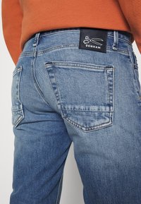 Světlé modré džínové kalhoty s dvěma zadními kapsami, černou štítkem s bílým logem na pasu a rovnými nohavicemi.