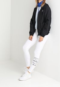 Kvinna i svart zip-up jacka, blå topp, vita leggings med flerspråkig text nära vaden, och vita sneakers, står mot en vit vägg.