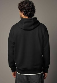 Homme avec des cheveux bouclés portant un sweat à capuche noir et un jean sombre, se tenant de dos contre un fond gris uni.