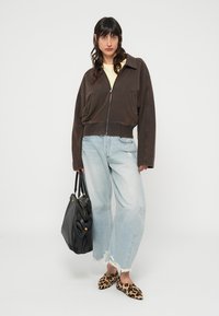 Veste marron courte à fermeture éclair, haut jaune clair, jean large bleu clair avec ourlets effilochés, sac à main noir et chaussures à enfiler imprimé léopard.