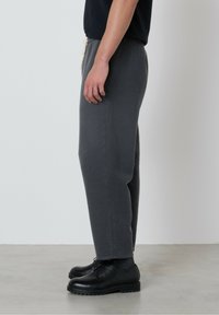 Osoba oblečená v tmavošedých teplákových nohaviciach, čiernych ponožkách, čiernych šnurovacích topánkach a čiernom tričku s krátkym rukávom, stojaca bokom na šedom podlaží.