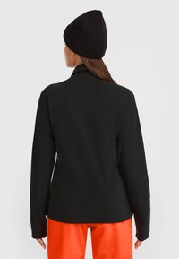 Svart fleecjacka med hög krage och långa ärmar. Slät textur, nedre fållen är rundad, bärs tillsammans med klara orangea byxor, vilket ger en avslappnad look.