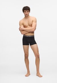 Czarne bokserki z białą, markową, elastyczną talią. Model ma odkryty tors i stoi boso, z założonymi rękami. Proste tło.