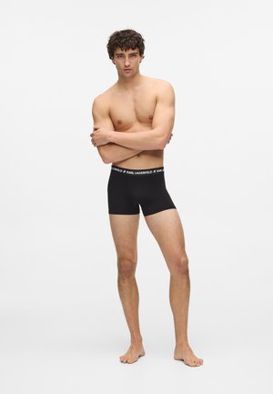 Boxer neri con una fascia elastica bianca brandizzata. Il modello ha il torso nudo e sta in piedi a piedi nudi, con le braccia incrociate. Sfondo semplice.