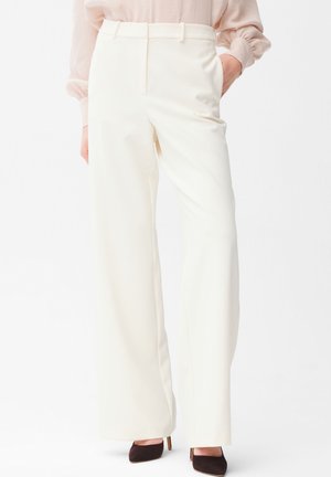 Frau trägt weiße, hoch taillierte, weit geschnittene Hosen, hellrosa Bluse und schwarze spitze High Heels, steht mit einer Hand in der Tasche.