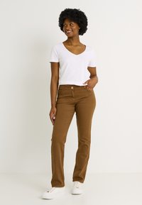 Pantalon marron à jambes droites avec poches avant et fermeture par bouton, associé à un t-shirt blanc à col en V et des baskets blanches. Texture lisse.