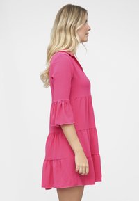 Vaaleanpunainen kerroksittain laskostettu mekko, jossa on kolme neljäsosahihat, tekstuuri kangas, liehukkahihansuut ja rento istuvuus. Mekossa on pystykaulus ja röyhelöinen helma.