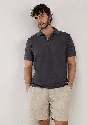 Hombre con el pelo corto y rizado, vestido con una camiseta polo gris oscuro y pantalones cortos beige, de pie con las manos en los bolsillos frente a un fondo liso.