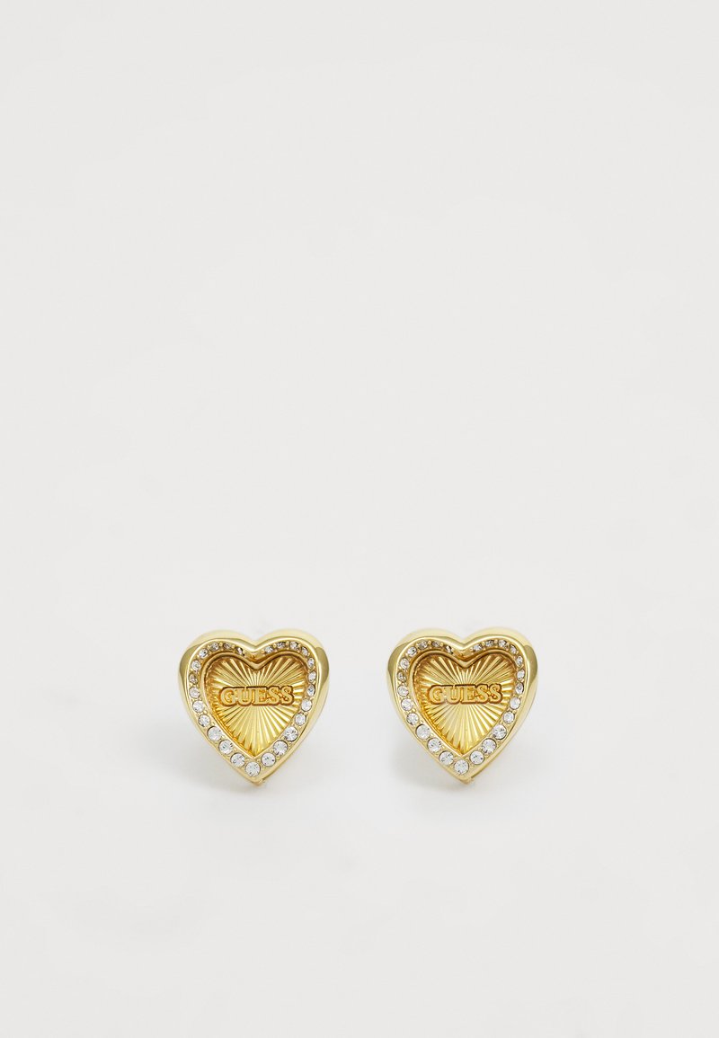 Zlatne naušnice u obliku srca s teksturiranim završetkom, koje sadrže "GUESS" logo i okružene su redom prozirnih rhinestones.