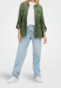 Giacca di tessuto verde con chiusura a bottoni, due tasche sul petto e maniche arrotolate, abbinata a jeans dritti blu chiaro e scarpe da ginnastica bianche.