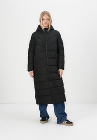 Zwarte pufferjas met capuchon, voorzien van een lange, gewatteerde uitstraling, verticale stiknaden en ritssluiting. Draag het over blauwe wijde jeans en sneakers.