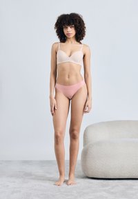 Σουτιέν σε ανοιχτό γυμνό χρώμα με ομαλή υφή, με λεπτές τιράντες, συνοδευόμενο από ροζ μπικίνι χωρίς ραφές, εμφανιζόμενο σε απαλό γκρι φόντο.