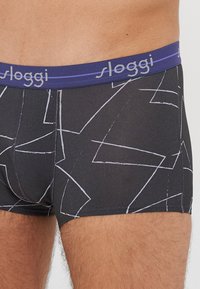 Black boxer briefs with a geometric white design, featuring a purple waistband with "sloggi" logo. Made from smooth, stretchy material.