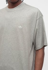 Άτομο που φοράει ένα ανοιχτό γκρι T-shirt της Adidas με μικρό λευκό λογότυπο Adidas trefoil στο αριστερό μέρος του στήθους.