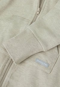 Hellbeige Zip-up Kapuzenjacke aus weichem Stoff, mit gerippten Bündchen, sichtbaren Nähten und einem kleinen grauen Etikett mit "Polarn O. Pyret."