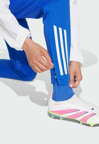 Blaue Sporthose mit weißen Streifen, ausgestattet mit Reißverschlüssen an den Bündchen. Der Träger kombiniert sie mit einem weißen und pinken Fußballschuh.