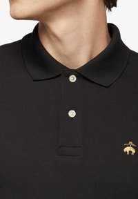 Černé polo tričko s texturovanou tkaninou, třemi krémovými knoflíky a malým zlatým zvířecím emblémem na levé části hrudi.