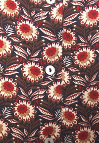 Tissu fleuri bleu marine avec de grandes fleurs beiges et rouges. Présente des motifs de feuilles détaillés et des accents de boutons blancs. Tissu en coton texturé.