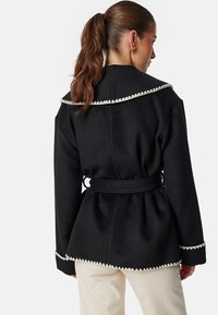 Czarna kurtka z szerokim kołnierzem, dekoracyjnym białym wykończeniem i strukturalną powierzchnią. Taperowany krój z ukośnymi rękawami.