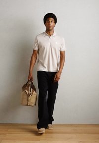 Beige színű galléros póló, fekete nadrág és bézs kézitáska barnás részletekkel. A modellen afro frizura van, és egy fával burkolt padlón áll.