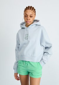 Cropped hoodie azul claro com um logotipo gráfico circular no peito, combinado com calções verdes. O hoodie possui um cordão e punhos canelados.