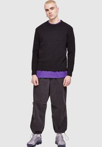 Pulover negru din tricot cu guler rotund peste o cămașă cu mânecă lungă violetă, asortat cu pantaloni gri lejeri și adidași gri.
