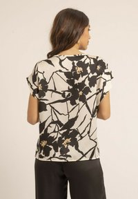 Blouse à manches courtes, crème avec un motif floral noir et des accents dorés. Texture lisse, coupe ample et ourlet arrondi.
