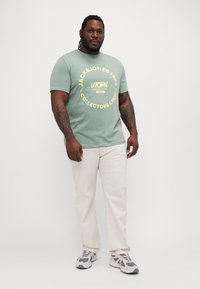 T-shirt verde chiaro con stampa grafica gialla, abbinato a jeans bianchi e sneakers grigie. Uomo in piedi, che mostra un completo casual.