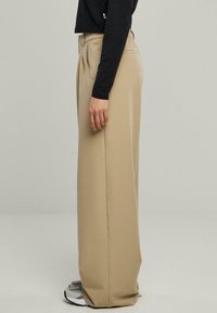 Un pantalon beige à jambes larges avec une taille haute, en matériau lisse et doté de poches latérales. Assorti avec un haut noir à manches longues et des baskets grises.