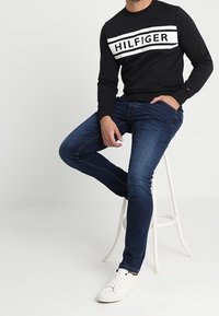 Sudadera negra con el logotipo blanco "HILFIGER", combinada con jeans azul oscuro. El modelo lleva zapatillas blancas y está sentado en un taburete.
