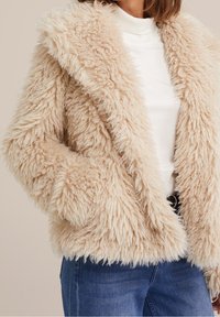 Beige fuskpälsjacka med en texturerad, fluffig yta, breda revers och långa ärmar, bärs över en vit tröja och blå jeans.