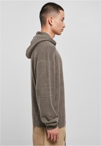 Gray knit hoodie with a relaxed fit, ribbed cuffs, and a large hood. Soft texture with a subtle sheen. Worn over beige pants.