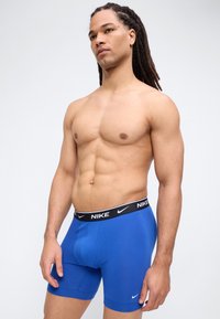 Μυώδης άνδρας με ντρέντλοκς, φορώντας μπλε μποξεράκια Nike, στέκεται μπροστά σε απλό, φωτεινό φόντο.