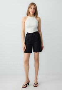 Witte geribbelde haltertop gecombineerd met hoge, gestructureerde zwarte shorts en zwarte hakslippers, tegen een lichte achtergrond.