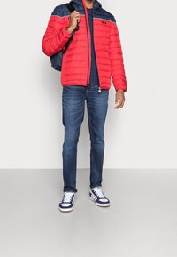 Rød pufferjakke med marineblå accenter, lynlåsåbning foran og hætte. Sammen med blå jeans og hvide sneakers med marineblå detaljer. Rygsæk på venstre skulder.