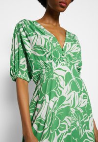 Zelené květinové šaty s bílými převislými květinami, hlubokým výstřihem do V, nafouknutými rukávy, elastickým pasem a dekoračními knoflíky vpředu.