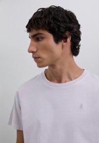 Joven con cabello rizado oscuro mirando a la izquierda, vistiendo una camiseta de cuello redondo ligera color púrpura liso con un pequeño logotipo de calavera en el pecho.